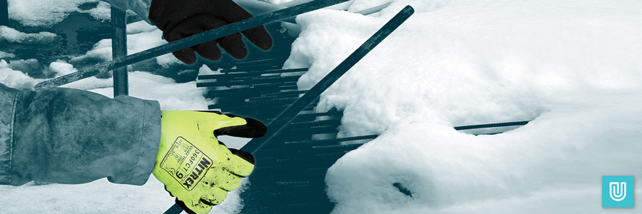 A construction worker wearing hi-vis Nitrex 360FCT safety gloves while handling metal rods on a construction site in very cold weather. The rods are covered in ice and snow.